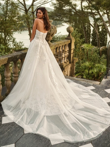 Bride wearing the Enzoani WALINA wedding dress featuring a strapless lace corset bodice, fitted floral lace silhouette and detachable tulle overskirt on an elegant lakeside terrace.