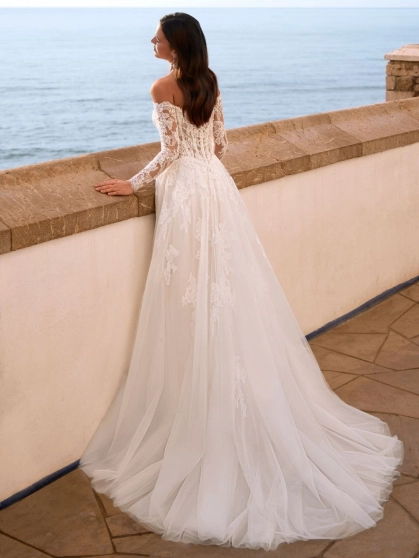 Love Faiza is a romantic A-line wedding dress from the 2027 collection featuring an illusion lace corset bodice, detachable off-the-shoulder long lace sleeves and a flowing soft tulle skirt with floral lace appliqués. Designed by Enzoani for brides seeking